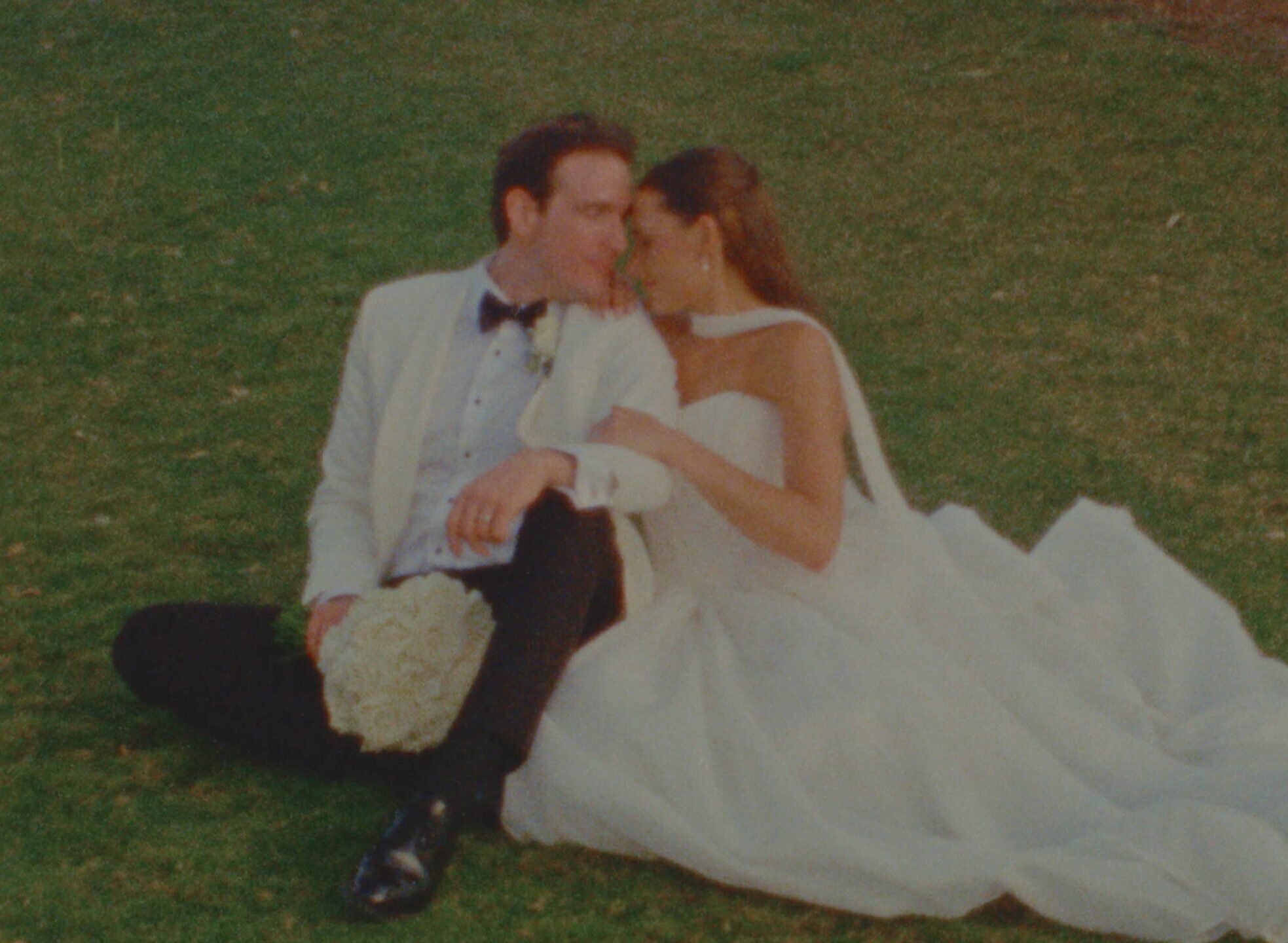 bride and groom seated in the grass with their foreheads touching